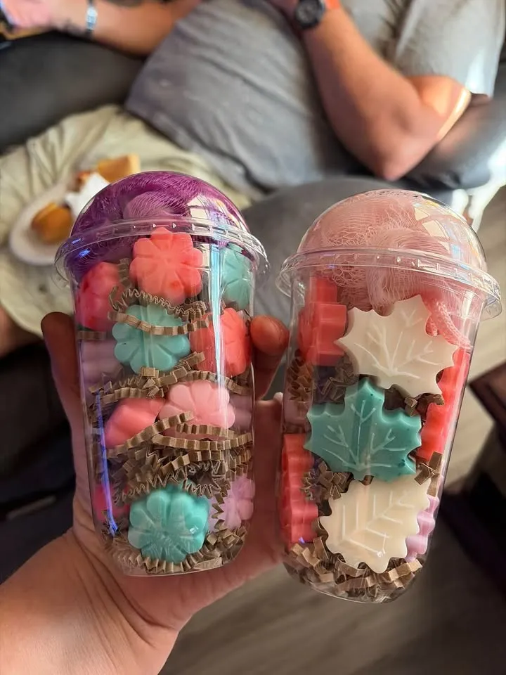 Two clear cups filled with flower and leaf shaped soaps, with loofah poufs on top.